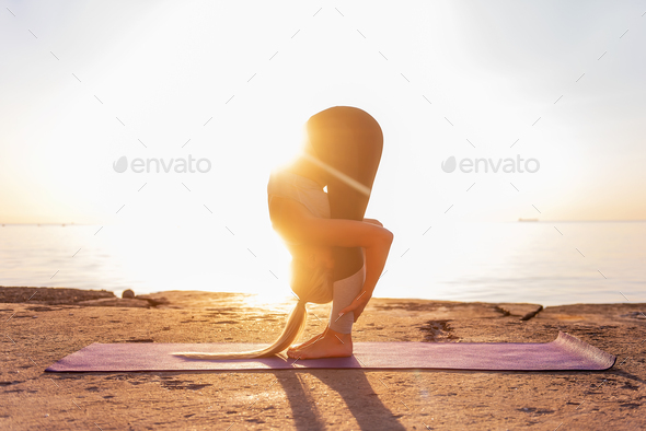 Young blonde woman in sportswear doing yoga asanas Uttanasana pose on the beach at sunrise