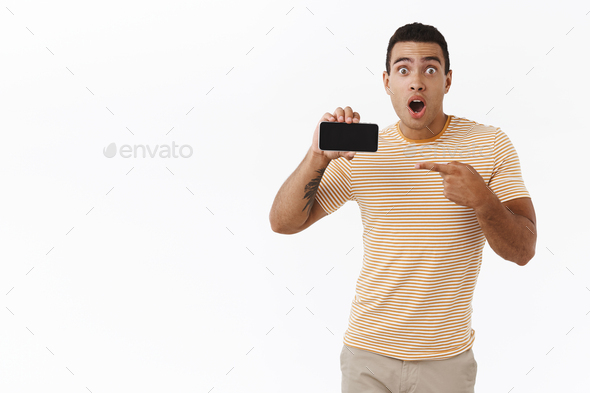 Amazed, speechless young masculine man, hold his breath as showing something wonderful on smartphone