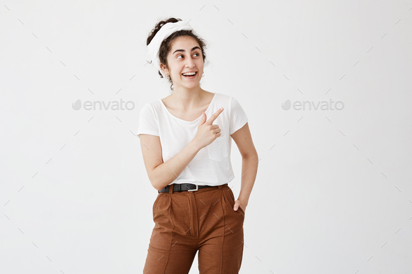 Excited young dark-haired woman with hair knot pointing her index finger sideways, raising eyebrows
