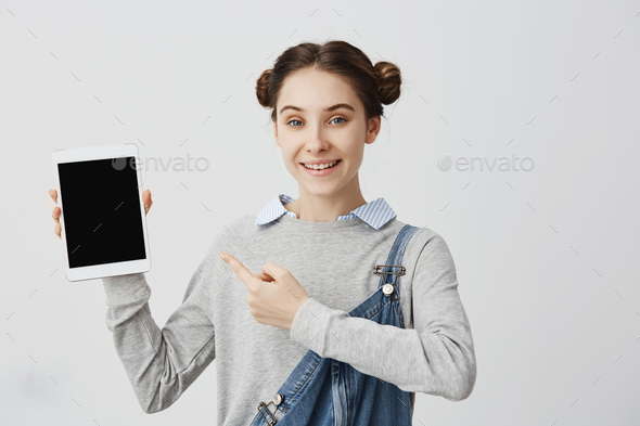Smiling adult girl with hair tied in double buns holding modern device pointing finger on it. Joyful