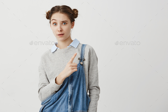 Caucasian woman 20s wondering about what she see showing index finger sideways raising eyebrow. Girl