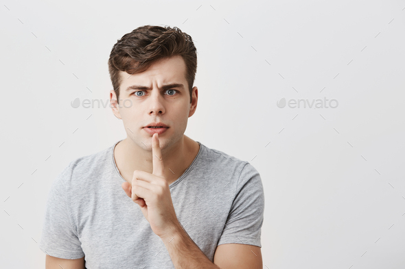 European male model in gray t-shirt keeping index finger on his lips, frowning his face, asking to