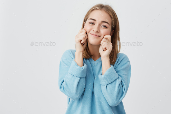 Cute funny attractive girl with straight fair hair pinching her cheeks and smiling, wearing blue