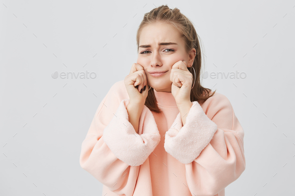 Isolated shot of pretty charming blonde caucasian girl in pink clothes pinching her cheeks, showing