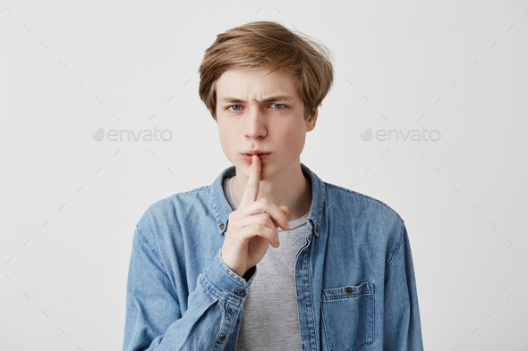 Attractive pleasant looking young man frowns face keeps finger on lips, asks friends not to tell