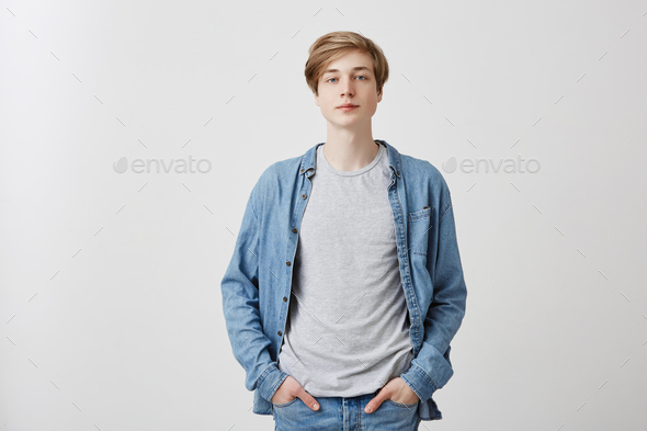 Portrait of pensive young fair-haired guy with blue eyes wears denim clothes, keeps hands in pockets