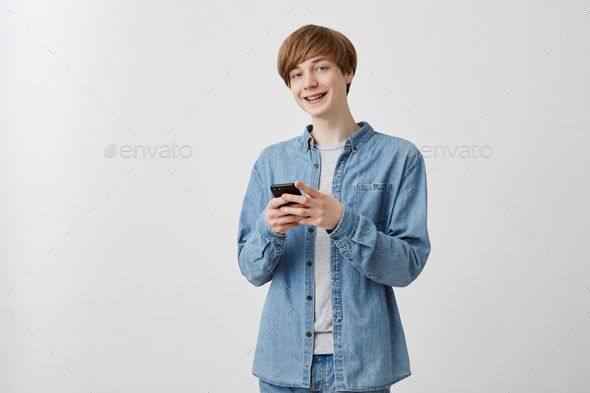 Cheerful male blogger with fair hair dressed in denim clothes, uses app on smart phone, enjoyes