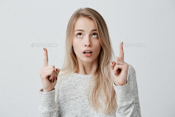 Blonde girl in sweater with astonished face expression keeping index fingers pointed upwards. Cute