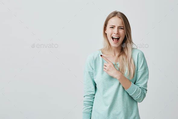 Amazed shocked female model with straight fair hair, wearing blue clothes, posing with widely opened