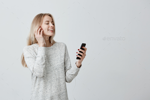 Glad attractive female with blonde dyed hair standing indoors with her smartphone chatting with