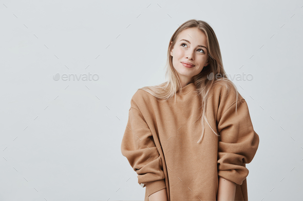 Pleased delightful smiling female with blonde dyed hair, has dreamy expression, wears cozy sweater