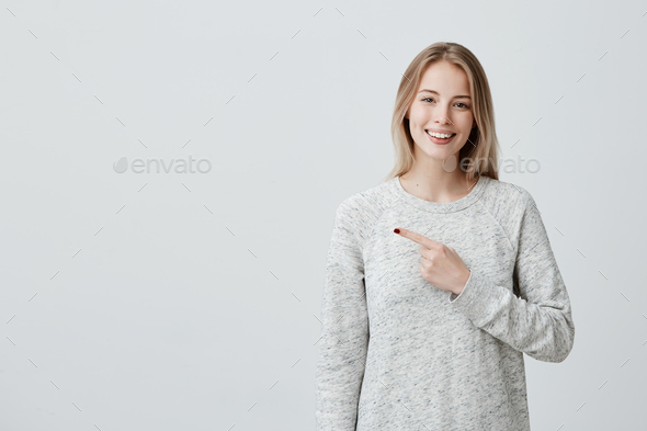 Smiling joyful woman with blonde dyed hair in loose sweater posing against gray studio wall pointing