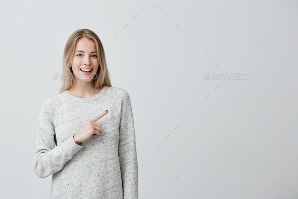 Joyful happy young blonde woman with dyed hair pointing with forefinger at wall with copy space for