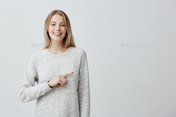 Happy joyful young blonde woman with dyed hair smiling broadly pointing with index finger at wall