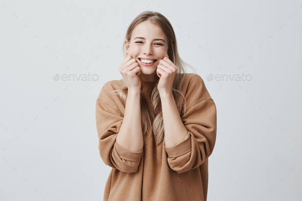 Charming beautiful female model smiling broadly wearing brown sweater, pinching her cheeks, mocking