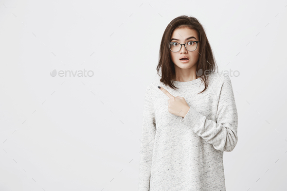 Portrait of caucasian cute brunette in glasses, lifting eyebrows in concern and shock, pointing