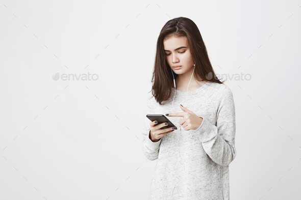 Young attractive european student listening music and scrolling news feed in her smartphone with
