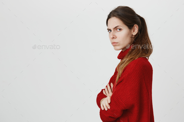Profile of beautiful girl with ponytail in warm red sweater posing at studio, keeping arms folded