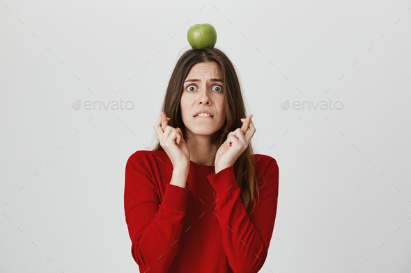 Stressed young woman in red sweater with green apple on head biting lower lip crossing fingers with