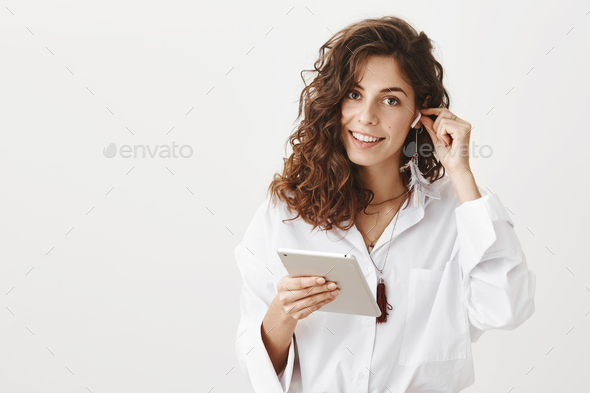 Portrait of feminine charming caucasian woman with curly hair inserting wireless earphone in ear