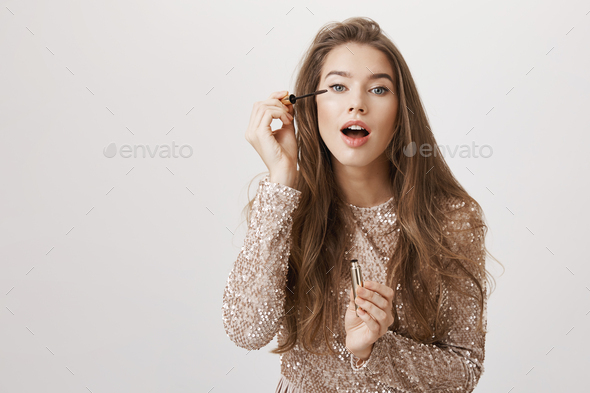 Pretty feminine european woman putting on mascara with opened mouth, wearing glittering trendy