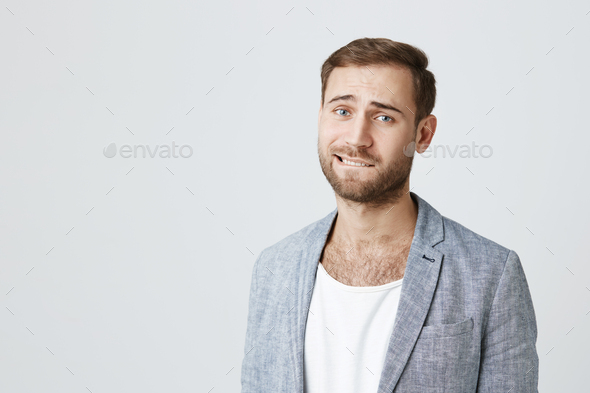 Good looking male model with stubble bites lower lip, looks pensively at camera, thinks over his