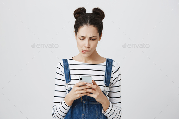 Shocked brunette female with hairbuns holds mobile phone, missed important call, being puzzled