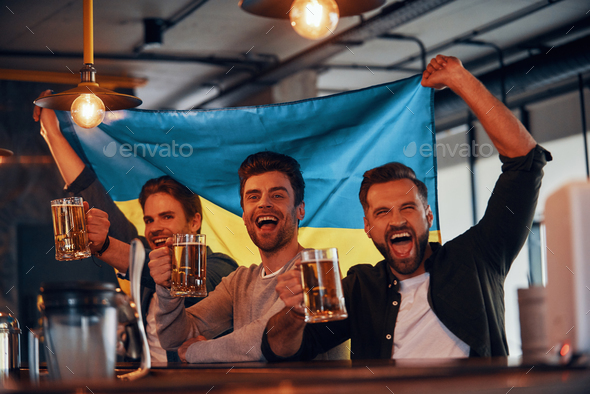 Three cheering young men carrying Ukrainian flag and enjoying beer