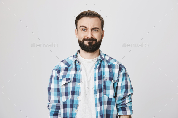 Portrait of funny disappointed bearded man, lifting his eyebrow and fake smiling while looking at