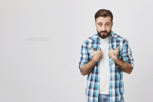 Studio shot of bearded european person pointing at himself with offended expression, standing