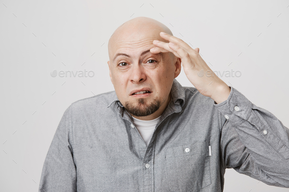 Studio portrait of bald mature european man touching eyebrow and expressing low spirits while