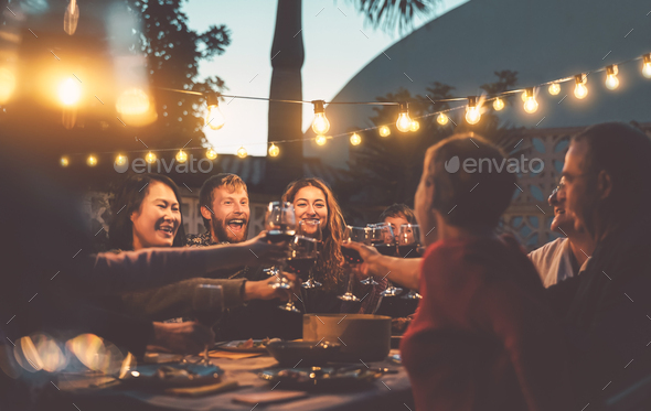 Young people celebrating together drinking red wine glasses on patio garden  at summer party. Diverse friends having fun cheering glasses during dinner  party. Friendship and celebration concept Stock Photo