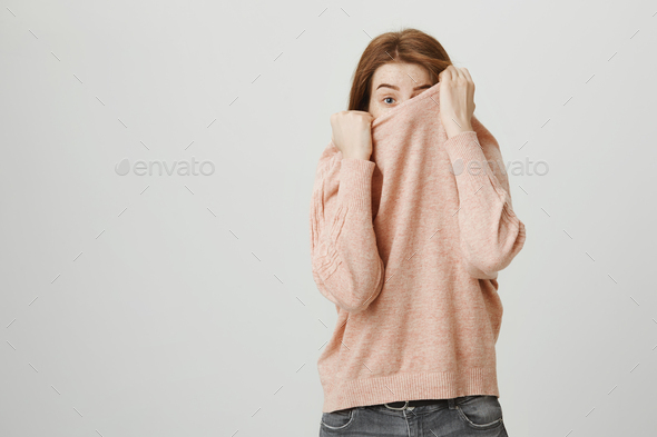 Studio portrait of funny european redhead woman hiding in her pullover while peeking with one eye