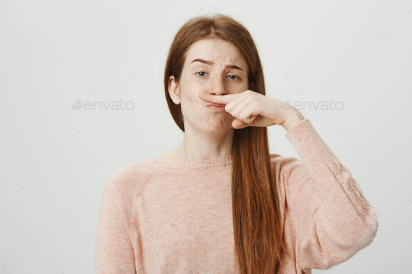 Portrait of funny upbeat ginger girl holding index finger above nose as if it is moustache and