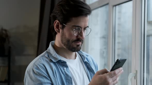 Young happy man standing near window indoors at home while using mobile phone alt
