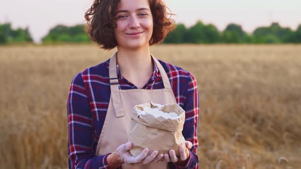 Joyful Smiling Woman With Flour. Woman With Hands Flour Holds Bag. Background Of Golden Wheat Field alt