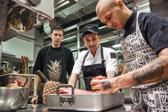 Professional chef in apron holding a red meat and teaching his assistants