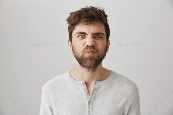 Portrait of funny weird guy with messy hair and beard making faces, puckering eyebrows and sulking