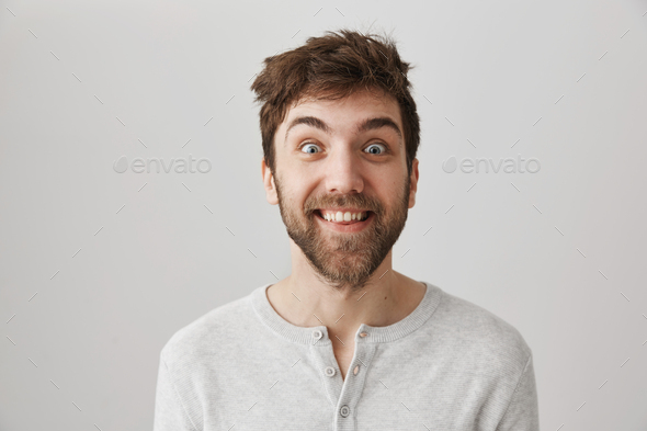 Funny and crazy bearded guy with messy hair and popped eyes, smiling and staring at camera, standing