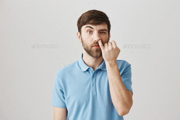 Portrait of funny impolite caucasian man with beard, picking nose with pinky staring with lifted