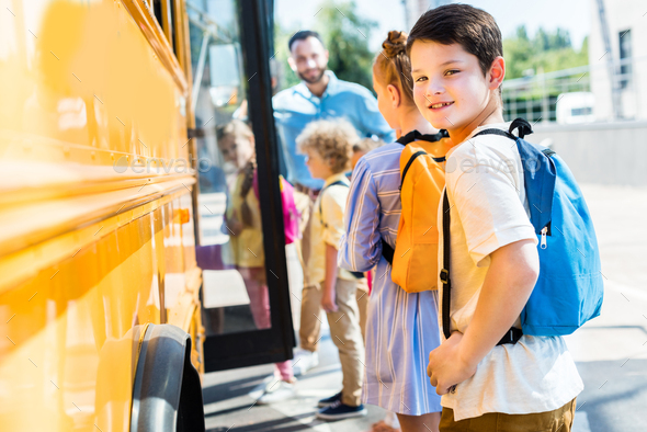 little schoolboy entering school bus with classmates while teacher standing near door