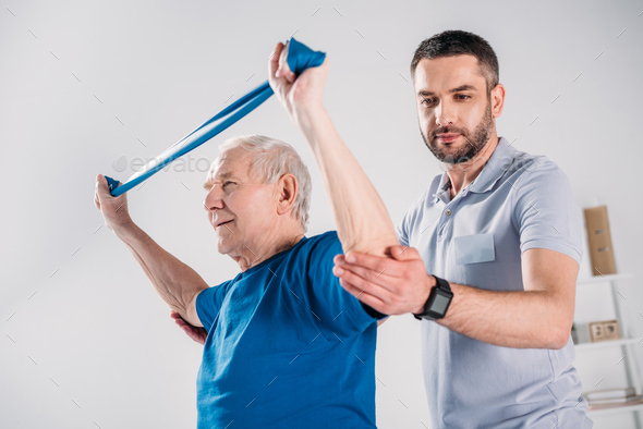 side view of rehabilitation therapist assisting senior man exercising with rubber tape