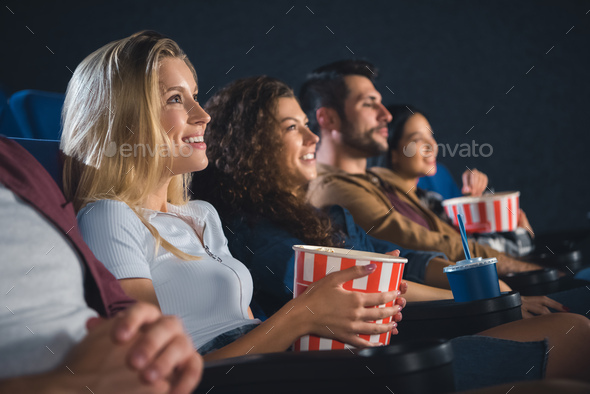 smiling multiethnic friends with popcorn watching film together in movie theater