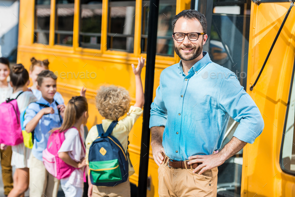 handsome bearded teacher looking at camera while pupils entering school bus blurred on background