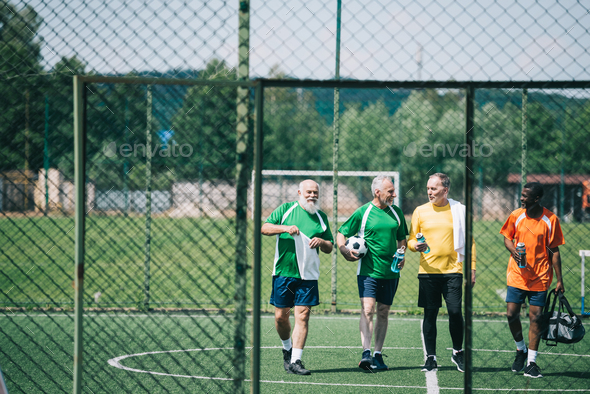group of interracial elderly sportsmen with sportive water bottles walking on football field