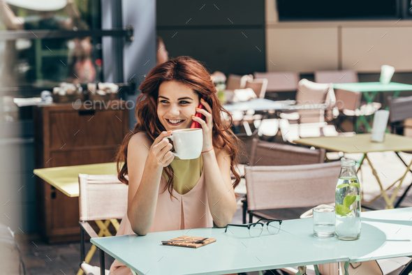 portrait of smiling woman with cup of coffee talking on smartphone in cafe