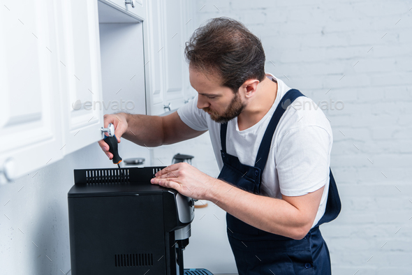 bearded handyman in working overall repairing coffee machine by screwdriver in kitchen