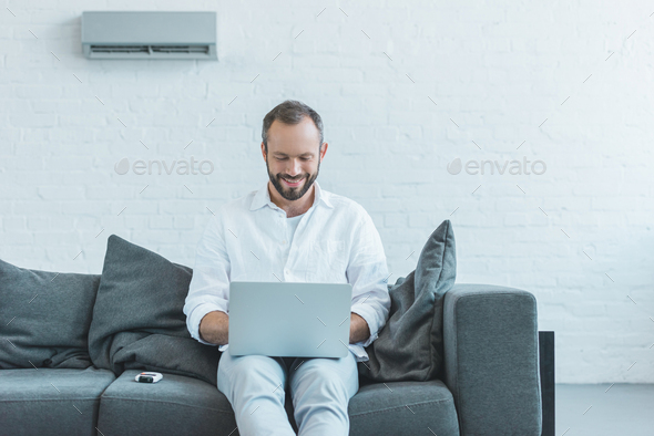 smiling man using laptop and sitting on sofa, with air conditioner on wall