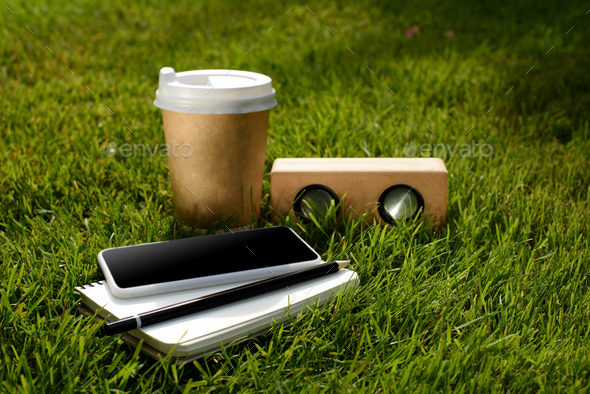 close up view of coffee to go, smartphone, notebook and audio speaker on green grass