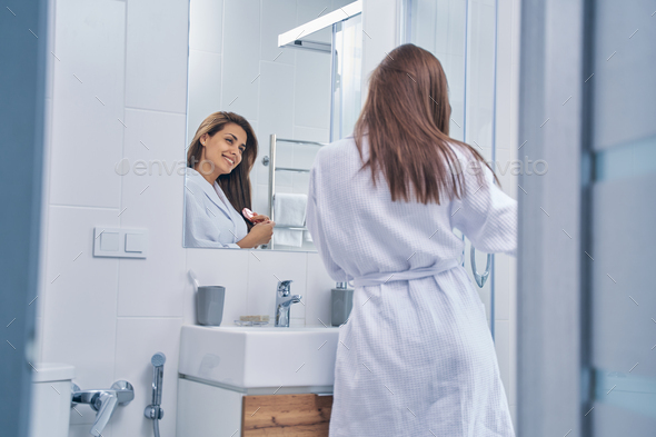 Cheerful young woman brushing hair in bathroom Stock Photo by Iakobchuk
